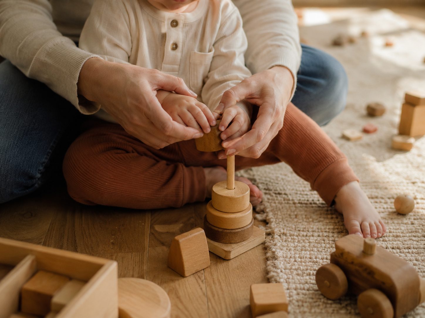 A parent's hands guiding a toddler stacking wooden toys on the floor